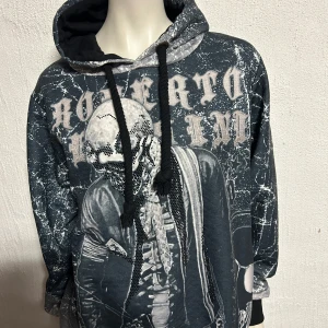 Svart hoodie med tryck från Roberto Cavalli - Cool svart hoodie från Roberto Cavalli med ett unikt tryck av en dödskalle och text på framsidan. Hoodien har en justerbar huva med snören och ett mönster som ger en sliten look. Perfekt för en edgy stil!