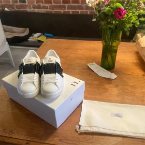 Valentino skor - Snygga vita Valentino sneakers med svarta detaljer och nitar. Skorna har en unik design med en bred rem över snörningen och en stilren siluett. Perfekta för att ge din outfit en lyxig touch.