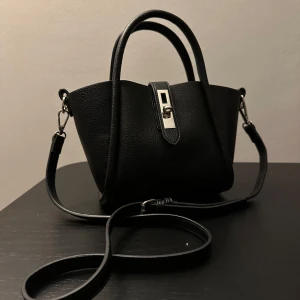 Svart axelväska med dragkedja - Snygg svart axelväska i skinn med praktisk dragkedja och justerbar axelrem. Väskan har ett stilrent spänne framtill och två handtag för enkel bärning. Perfekt för att hålla dina saker säkra och organiserade.