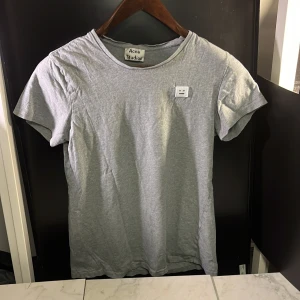 Grå t-shirt från Acne Studios - Säljer en stilren grå t-shirt från Acne Studios. Den har en klassisk rund hals och korta ärmar. Perfekt för en avslappnad look, bra att veta att den sitter lite annorlunda i axlarna. Det finns även defekter som syns på bild nr.4, hör av er vid minsta fundering🙌