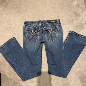 Blå jeans från Rock Revival  - As coola blå bootcut jeans från Rock Revival i strl.27 med broderade detaljer på bakfickorna. I bra skick och sparsamt använda, saknas dekoration på en av knapparna men inget man lägger märke till ❣️ 