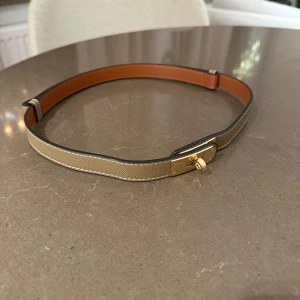 Beige bälte med gulddetaljer - Säljer ett stilrent beige bälte med guldfärgat spänne. Perfekt för att ge en elegant touch till din outfit. Bältet är i skinn och har en smal design som passar både till vardags och fest. Justerbar storlek för bästa passform.