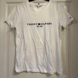 Vit t-shirt från Tommy Hilfiger - Säljer en klassisk vit t-shirt från Tommy Hilfiger med tryck. Den är kortärmad och i bomull, perfekt för en avslappnad stil. T-shirten har en liten logga på ärmen också. Den är knappt använd. Ett litet hål uppe vid nacken som syns på bild, lätt att laga.