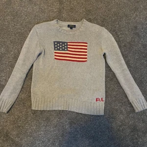 Grå stickad tröja från Ralph Lauren - Säljer en snygg grå stickad tröja från Ralph Lauren med en amerikansk flagga på bröstet. Tröjan har långa ärmar och ribbstickade muddar. Perfekt för höst och vinter! Vill få såld snabbt då därför lågt pris 