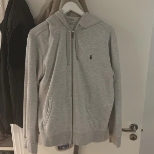 Grå hoodie från Polo Ralph Lauren - Säljer en snygg grå hoodie från Polo Ralph Lauren i storlek M. Den har en dragkedja framtill och en klassisk logga på bröstet. Perfekt för en avslappnad stil och passar till de flesta tillfällen. Huvan har dragsko och det finns fickor på sidorna. Superbekväm och stilren!