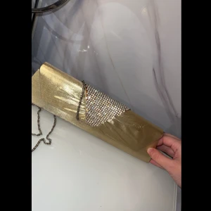 Guldig kuvertväska med paljetter - Snygg guldig kuvertväska perfekt för fest! Väskan har en glittrig paljett-detalj på locket och en elegant kedja som axelrem. Den är i syntetmaterial och ger en lyxig känsla. Perfekt för att lysa upp vilken outfit som helst!