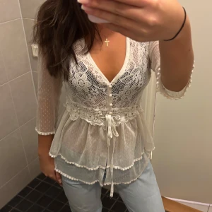 Vit spetsblus med knytband - Säljer en superfin vit spetsblus med trekvartsärm och v-ringning. Blusen har knappar framtill och ett sött knytband i midjan. Perfekt för vår och sommar, och funkar både till vardags och fest. Den har ett vackert blommönster och volangdetaljer som ger en romantisk touch. 💕💕 Storlek L men passar mig bra som har xs-s/34!