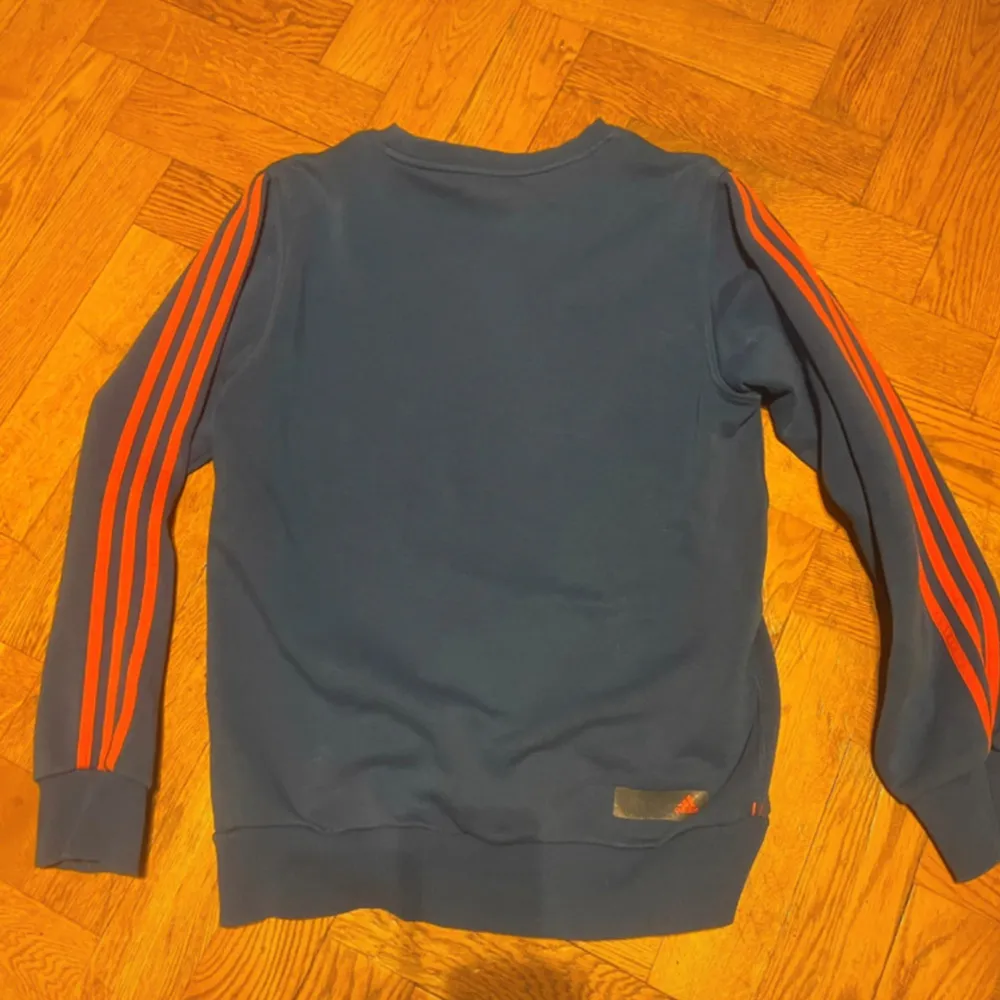 Säljer en snygg blå tröja från Adidas med de klassiska tre ränderna i rött längs ärmarna. Tröjan har en rund halsringning och är perfekt för en sportig look. Den är i bra skick och passar perfekt för höst och vår.. Hoodies.