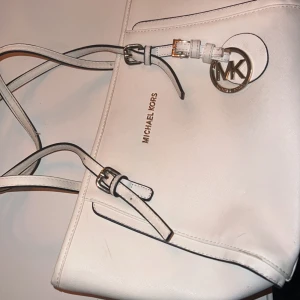 Vit axelväska från Michael Kors - Säljer en stilren vit axelväska från Michael Kors i mycket bra skick. Väskan har en elegant design med ett stort MK-emblem och justerbara remmar. Insidan är rymlig med flera fack och dragkedja för säker förvaring. Perfekt för både vardag och fest!
