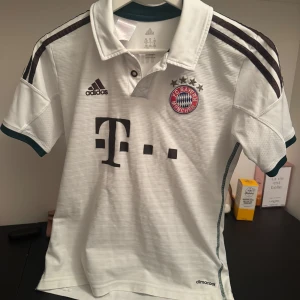 Vit Bayern München fotbollströja från Adidas - Säljer en vit Bayern München fotbollströja från Adidas i bra skick. Tröjan har korta ärmar och klassisk krage med knappar. Den har klubbens emblem på bröstet och Adidas-loggan. Perfekt för fans av laget! ⚽
