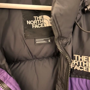 Jacka från The North Face - Säljer en snygg lila dunjacka från The North Face i storlek M. Jackan har en svart axelparti och dragkedja framtill. Perfekt för kalla vinterdagar med sin varma och puffiga design. Den här tyvärr lite trasig dragkedja men självklart är det något som går o fixa🥰
