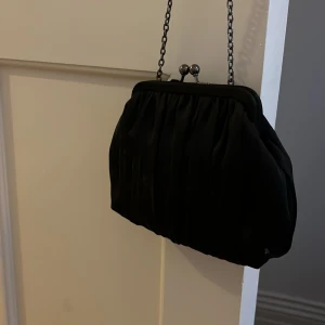 Svart kuvertväska med kedja - Säljer en elegant svart kuvertväska med en stilren kedja. Väskan har en klassisk design med en metallknäppning upptill och är perfekt för festliga tillfällen. Insidan är fodrad med ett ljust tyg och har en liten ficka för extra förvaring. Perfekt accessoar för en kväll ute! Kom gärna med prisförslag ☺️