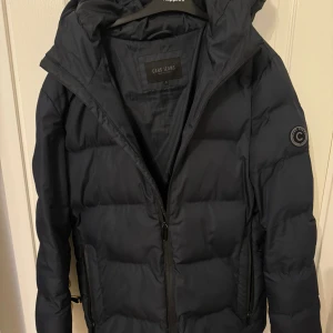 Mörkblå dunjacka från Cars Jeans - Säljer en snygg mörkblå dunjacka från Cars Jeans. Jackan har en dragkedja framtill och en mysig huva. Perfekt för kalla vinterdagar. Den är i mycket bra skick och har en stilren design med ett märke på ärmen. Passar både till vardags och lite finare tillfällen.