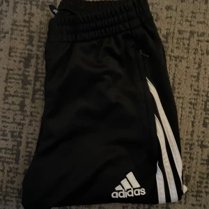 Svarta träningsbyxor från Adidas - Säljer ett par svarta träningsbyxor från Adidas. De har de klassiska vita ränderna längs sidorna och en elastisk midja för bästa komfort. Perfekta för träning eller en avslappnad dag. Passar både höst och vår.