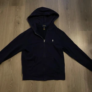 Mörkblå zip hoodie från Ralph Lauren. - Säljer en stilren mörkblå zip hoodie från Ralph Lauren i storlek L i barn. Den har en dragkedja framtill och en liten broderad logga på bröstet. Perfekt för både höst och vinter, och passar till alla tillfällen. Superbekväm och i bra skick! Pris kan diskuteras.