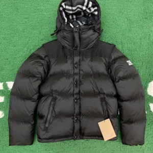 Svart dunjacka från Burberry - Säljer en snygg svart dunjacka från Burberry i nytt skick. Jackan har en avtagbar huva med klassiskt Burberry-mönster på insidan. Den är perfekt för vintern med sin varma vaddering och stilrena design. Jackan har knappar och dragkedja framtill samt praktiska fickor.