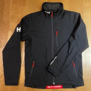 Helly Hansen jacka  - Säljer en marinblå Helly Hansen jacka i mycket bra skick. Den har en normal passform och är perfekt för höst och vår. Jackan har långa ärmar och praktiska dragkedjor i rött. Materialet är syntetiskt och håller dig varm och torr. Perfekt för utomhusaktiviteter!