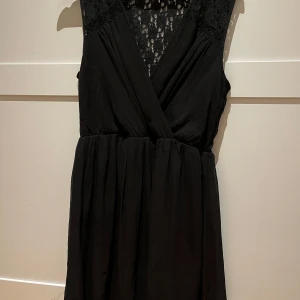 Svart klänning med spetsdetaljer - Elegant svart klänning från Vero Moda med v-ringad omlott-design och spetsdetaljer på axlar och rygg. Perfekt för festliga tillfällen. Klänningen är ärmlös och har en normal passform som ger en smickrande siluett.