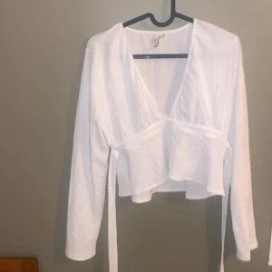 Vit omlottopp med knytband - Säljer en superfin vit omlottopp med långa ärmar och knytband i midjan. Perfekt för både vardag och fest! Toppen har en v-ringad design och är i mycket bra skick. Passar perfekt till jeans eller kjol för en chic look. I storlek S