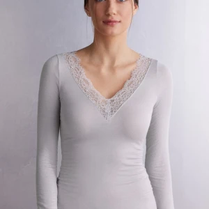 Vit långärmad topp med spetsdetaljer - Säljer en superfin grå långärmad topp från intimissimi med v-ringning och spetsdetaljer runt halsen. Den är i ett mjukt och stretchigt material som sitter bekvämt på kroppen. Använd fåtal gånger