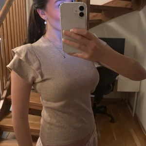 Beige topp med volangdetaljer - Säljer en superfin beige topp med volangdetaljer på ärmarna. Den är tight och perfekt för både vardag och fest. Materialet känns mjukt och bekvämt, och den har en stilren design som passar till det mesta. Perfekt för vår och sommar!