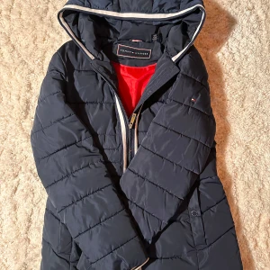 Navyblå pufferjacka från Tommy Hilfiger.  - Säljer en snygg navyblå pufferjacka från Tommy Hilfiger. Jackan har en dragkedja och en huva med stiliga detaljer. Perfekt för kyliga dagar och håller dig varm och bekväm. Den är i mycket bra skick och har en röd insida som ger en cool kontrast.