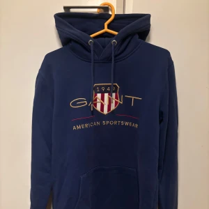Mörkblå hoodie från GANT - Säljer en snygg mörkblå hoodie från GANT med deras klassiska logga och texten 'American Sportswear' på framsidan. Perfekt för en avslappnad stil eller kyliga dagar. Nypris ligger runt 1500kr. Priset är inte hugget i sten🪨Skriv om du har npgra funderningar😉