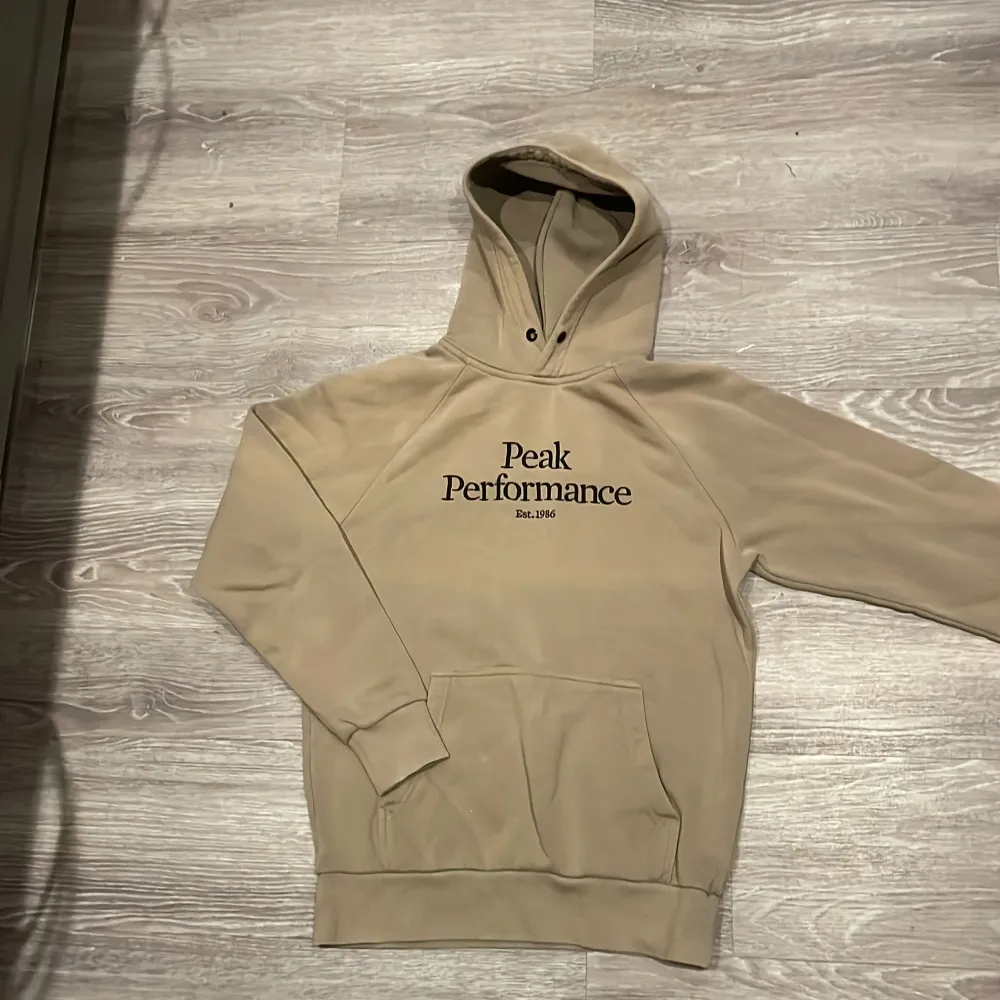 Säljer en snygg beige hoodie från Peak Performance. Den har en klassisk design med en stor ficka fram och justerbar huva. Perfekt för både vardag och chill. Passar till allt och är superbekväm!. Tröjor & Koftor.