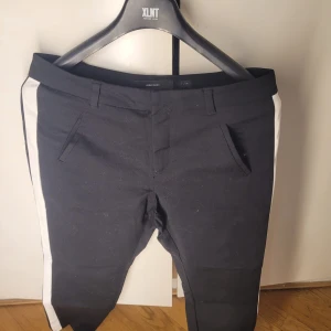 Svarta byxor med vita ränder från Vero Moda - Snygga svarta byxor från Vero Moda i storlek L. De har vita ränder längs sidorna som ger en sportig look. Byxorna har en normal passform och är perfekta för både vardag och fest. De är i mycket bra skick och redo för nya äventyr!