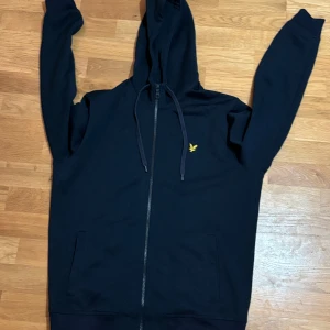 Mörkblå hoodie från Lyle & Scott - Säljer en snygg mörkblå hoodie från Lyle & Scott i storlek L men passar även M. Den har en dragkedja framtill och en liten gul logga på bröstet. Perfekt för höst och vinter, och den är superbekväm med långa ärmar och en praktisk huva. Passar både till vardags och chillkvällar. 🧢