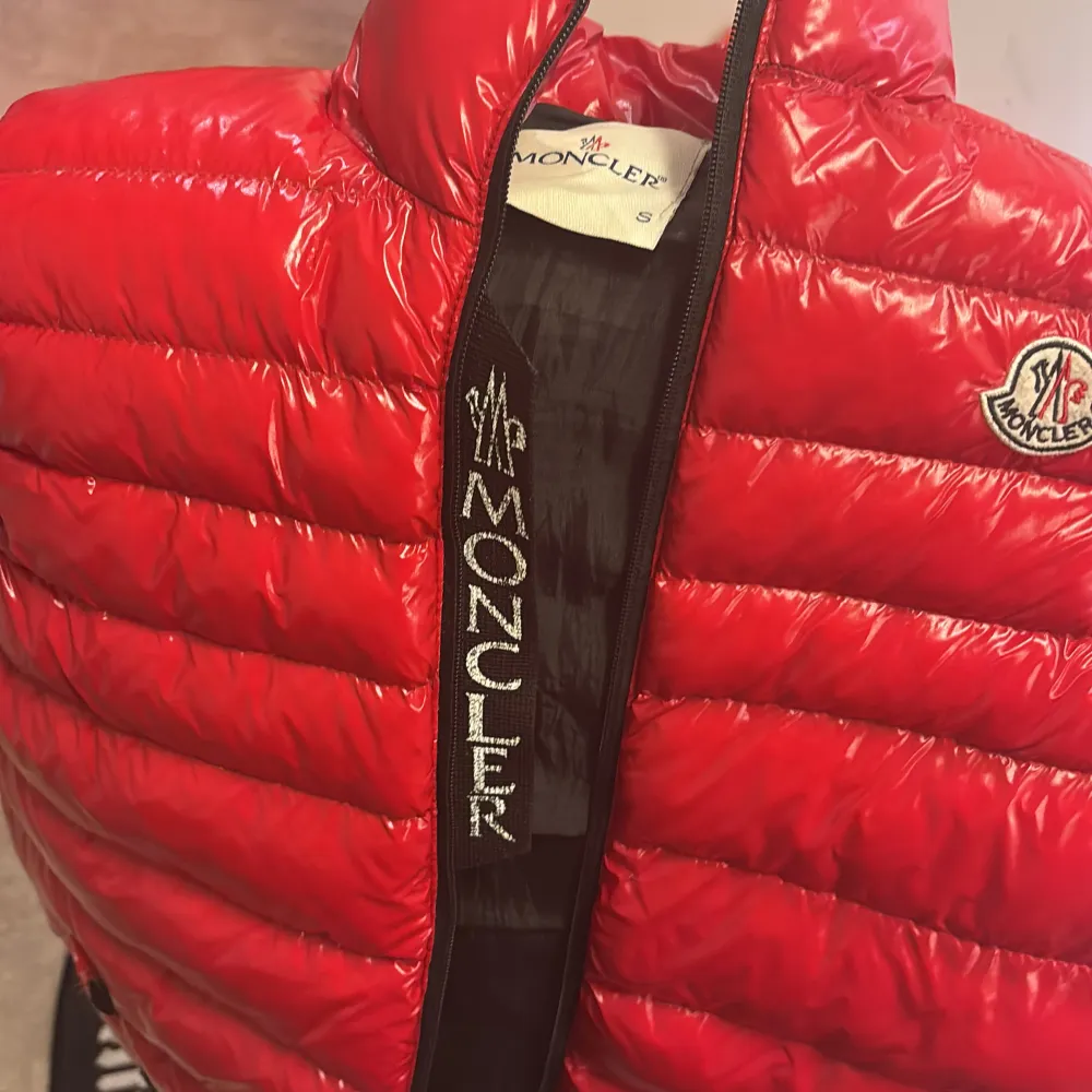 Säljer en snygg röd dunväst från Moncler i storlek S. Västen har en glansig finish med dragkedja och Moncler-logga på bröstet. Perfekt för kyliga dagar och ger en stilren look. Inuti finns en rolig serietidningsdetalj. Passar både höst och vinter.. Jackor.