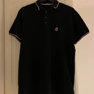 Svart pikétröja från Moncler - Säljer en svart pikétröja från Moncler. Den har korta ärmar och en krage med röd och vit rand. Tröjan är i bra skick! Priset kan förhandlas.