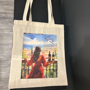 Beige tygg shoppingväska med eget tryck - Säljer en beige shoppingväska med ett eget färgglatt tryck av en kvinna i röd klänning som står på en balkong med utsikt över en pittoresk stad. Texten 'World's best mom' är tryckt i hörnet. Perfekt för vardagsbruk eller som present till mamma!