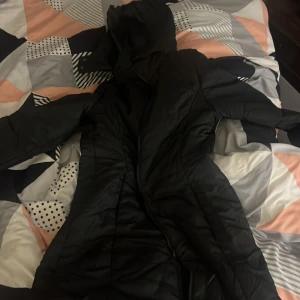 Svart lång dunjacka - Säljer en svart lång dunjacka i mycket bra skick. Den är perfekt för kalla vinterdagar och har en skön huva. Jackan har en dragkedja framtill och är quiltad för extra värme. Passar perfekt för vinterns alla äventyr!