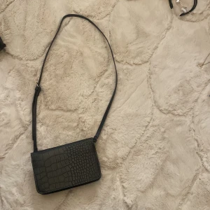 Svart axelväska från H&M - Snygg svart axelväska från H&M i krokodilpräglat mönster. Väskan har en justerbar axelrem och stängs med dragkedja. Perfekt för både vardag och fest! 🖤