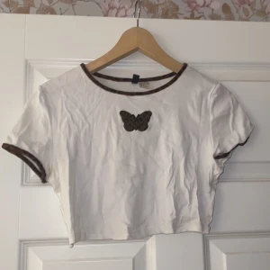 Vit croppad topp med fjärilsmotiv från Divided - Säljer en söt vit croppad topp från H&M Divided med ett brunt fjärilsmotiv på bröstet. Toppen har korta ärmar och bruna kanter vid hals och ärmslut. Perfekt för sommardagar eller en avslappnad stil. 🦋Passar M och L