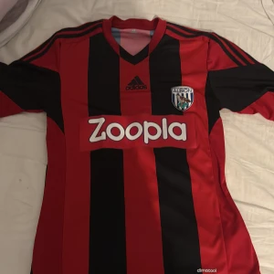 Röd och svart randig west bromwich från Adidas - Säljer en snygg röd och svart randig fotbollströja från Adidas med korta ärmar. Den har en V-ringning och klubbmärket på bröstet. Perfekt för fotbollsfans som vill ha en cool look på matchdagen! 🏆märket i mitten har blivit lite slitet och fått lite bubblor men funkar bra ändå!
