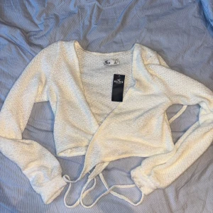 Vit omlottopp från Hollister - Säljer en superfin vit omlottopp från Hollister. Den är croppad och har långa ärmar, perfekt för en stilren look. Materialet känns mjukt och bekvämt, och den har knytband för att justera passformen. Passar perfekt till både vardag och fest!