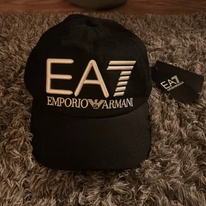 Svart keps från Emporio Armani - Säljer en snygg svart keps från Emporio Armani med den ikoniska EA7-loggan broderad framtill. Kepsen är i nyskick och har en justerbar rem baktill för perfekt passform. Aldrig använd. 