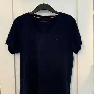 Säljer en snygg mörkblå t-shirt från Tommy Hilfiger. Den har en klassisk v-ringning och korta ärmar. Perfekt för en avslappnad stil och passar till det mesta. Den lilla loggan på bröstet ger en stilren touch.