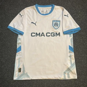 Vit fotbollströja från Puma - Säljer en vit fotbollströja från Puma med ljusblå detaljer och klubbmärke på bröstet. Tröjan har korta ärmar och rund halsringning. 