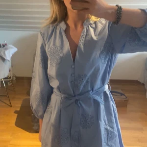 Ljusblå klänning med knytband - Säljer en ljusblå klänning med v-ringning och knytband i midjan. Klänningen har trekvartsärmar och ett fint broderat mönster. Perfekt för vår och sommar!