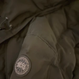 Svart dunjacka från Canada Goose - Säljer en svart dunjacka från Canada Goose med pälskantad huva. Jackan har en quiltad design och långa ärmar. Perfekt för kalla vinterdagar. Modellen är Wyndham. Storlek S, passa väldigt bra och känns bra vid användning, använd under denna vinter. Han som jag köpte av påstod att vara äkta, alltså är inte själv säker om äktheten!