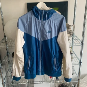 Nike windrunner - Säljer en snygg blå och vit vindjacka från Nike med dragkedja och huva. Jackan har en cool färgblockdesign med ljusblå, mörkblå och vita partier. Perfekt för blåsiga dagar med sin lätta och bekväma passform.