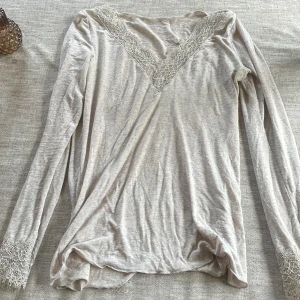 Beige långärmad topp från intimissimi - Säljer en stilren beige långärmad topp med v-ringad spetsdetalj vid halsen och ärmsluten. Perfekt för en elegant och avslappnad look. Passar bra till jeans eller kjol.