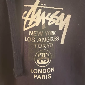 Stüssy X Dover Street Market - Säljer en svart hoodie som kan användas som jacka från Stüssy med tryck av olika städer som New York, Los Angeles och Tokyo. Hoodien har dragkedja och långa ärmar, perfekt för en casual look. Stussy i collaboration med Dover Street Market