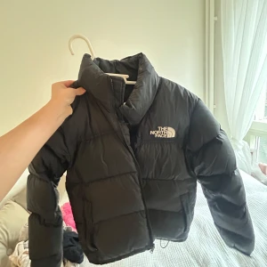 Svart dunjacka från The North Face - Säljer en svart dunjacka från The North Face. Jackan har en pufferdesign med dragkedja och är perfekt för kyligare väder. Den är långärmad och har en klassisk logga på bröstet. Perfekt för att hålla dig varm och stilren. Ursprungs pris var 3600