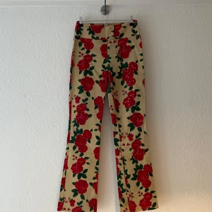 Blommiga byxor med rosor - Snygga vintage byxor med ett iögonfallande blommönster av röda rosor på en beige bakgrund. Byxorna har en bootcut-stil som ger en trendig look. Perfekta för att sticka ut i mängden!
