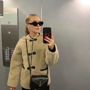 Beige teddyjacka med svarta detaljer - Säljer en mysig beige teddyjacka med svarta knappar och kantdetaljer. Jackan har en rund hals och långa ärmar, perfekt för kyligare dagar. Den har en stilren design med en twist av kontrasterande detaljer. Passar perfekt för en trendig look!