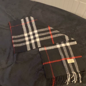 Rutig halsduk från Burberry 100% CASHMERE - Snygg rutig halsduk från Burberry i klassiska färger. Den har ett stilrent mönster med svarta, vita och röda rutor. Perfekt för att hålla dig varm och trendig under kyliga dagar. Fransen i ändarna ger en extra touch av stil.